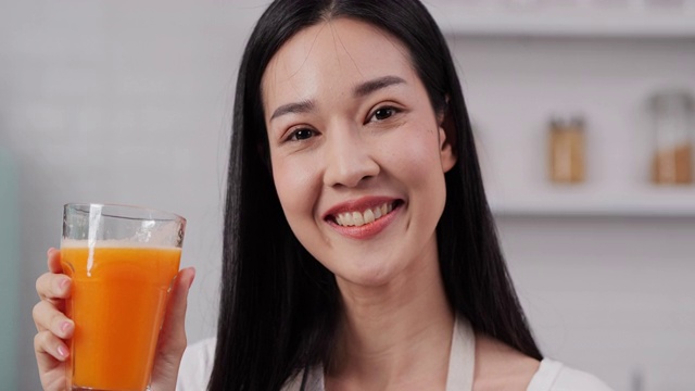 年轻的亚洲女子喝着橙汁，拿着一杯橙汁，微笑着看着镜头。