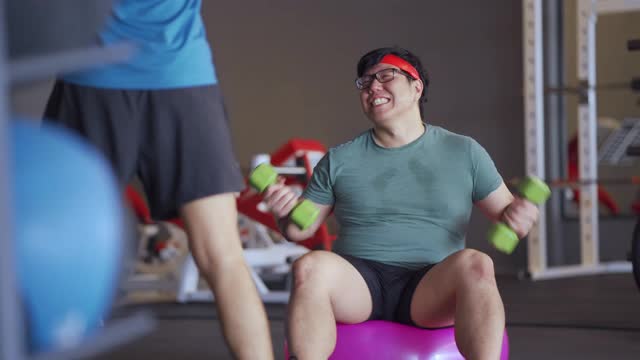 亚洲肥胖男性正在健身馆里用健身球上的哑铃努力工作，喜剧演员的概念