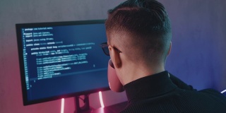 后视图的人程序员在眼镜编码网络安全软件室内霓虹灯。黑客晚上在漆黑的办公室里在电脑屏幕上输入代码