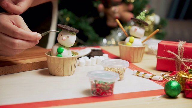 在装饰着圣诞树和装饰品的厨房里，准备冬天传统节日圣诞节的烘焙食品和甜点架，用糖霜自制纸杯蛋糕做一个雪人，用镊子给眼睛加巧克力