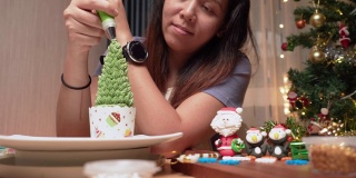 低角度的亚洲妇女准备烘焙糕点食品和甜点站为冬天的传统节日，自制纸杯蛋糕作为圣诞树，添加绿色奶油作为树叶子在桌子上装饰圣诞老人和企鹅糖衣。
