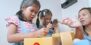 亚洲女孩正在把地址写进盒子里，以帮助父母为在线客户准备送货箱，而母亲使用笔记本电脑接收和检查网上购买的购物订单。