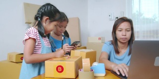亚洲女孩正在把地址写进盒子里，以帮助父母为在线客户准备送货箱，而母亲使用笔记本电脑接收和检查网上购买的购物订单。