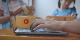 女人在笔记本电脑上打字，接收和检查网上购物订单。孩子正在把地址写进盒子里，帮助家长为在线客户准备快递盒。