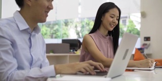 两个年轻的亚洲女商人和商人与笔记本电脑在办公大楼合作。20多岁的同事闲聊，头脑风暴。