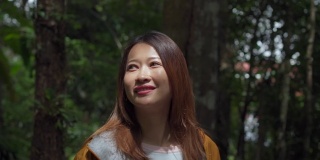 4K微笑的年轻美丽的亚洲女游客，穿着黄色夹克独自走在森林小径上。漂亮女孩徒步旅行者喜欢在美丽的大自然中徒步旅行。户外生活方式和旅游度假概念。