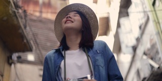 微笑的年轻亚洲女性旅行者戴着复古软呢帽走在泰国曼谷的一条小街道上，抬头环顾四周。