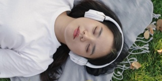 美丽的年轻亚洲女性戴着太阳镜和耳机睡在草地上。