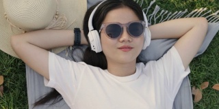 美丽的年轻亚洲女人戴着太阳镜和耳机睡在草地上。