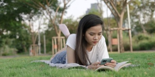 迷人的美丽的年轻亚洲女子放松阅读和使用智能手机，而躺在地板上的草地在一个公园的户外美丽的日落。