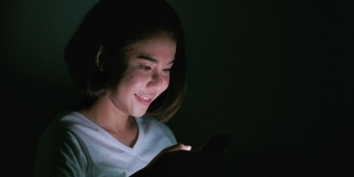 年轻的微笑女孩使用她的手机晚上躺在床上。
