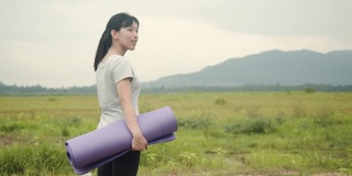 迷人的亚洲女人站着，握着瑜伽垫在美丽的山景在早上。
