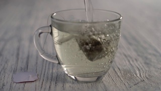 热水倒进一个玻璃杯子里，杯子里放着一袋凉茶，放在一张灰色的木制桌子上。视频素材模板下载
