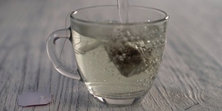 热水倒进一个玻璃杯子里，杯子里放着一袋凉茶，放在一张灰色的木制桌子上。