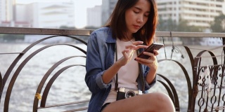 亚洲女性使用智能手机与她的朋友聊天，而坐在湄南河在泰国在美丽的日落。