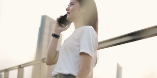 商务亚洲女人在一个白色的t恤是与朋友通电话，而阳光日落在现代办公大楼附近。