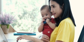 美丽的商业亚洲妈妈使用笔记本电脑和微笑，而花时间与她可爱的宝宝在家里。年轻的母亲在产假试图自由工作与幼童的书桌。