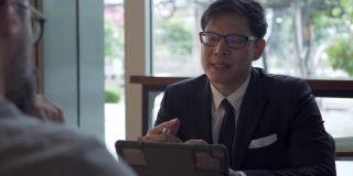 4K快乐的亚洲商人坐在咖啡店一起与白种人胡须男子创意和讨论商业计划与使用数字平板电脑。商务会议团队合作和技术理念