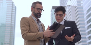 两个商务人士白种人与亚洲人同事伙伴关系讨论商业项目计划与使用数字平板电脑在城市。商务会议、技术、并购概念。