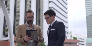 两个商务人士白种人与亚洲人同事伙伴关系讨论商业项目计划与使用数字平板电脑在城市。商务会议、技术、并购概念。