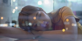 4K孤独年轻美丽的亚洲女人坐在窗前，躺在桌子上，晚上在咖啡馆用智能手机上网。悲伤的少女望着窗外灯火通明的城市街道