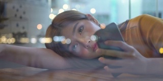4K孤独年轻美丽的亚洲女人坐在窗前，躺在桌子上，晚上在咖啡馆用智能手机上网。悲伤的少女望着窗外灯火通明的城市街道