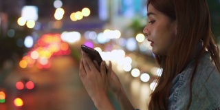 年轻美丽的亚洲女孩在夜晚走在城市里，喜欢用手机应用程序的智能手机相机拍下照片或记录下夜晚灯光照亮的城市景观的视频。