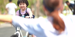 亚洲小女孩学习骑自行车与年轻的母亲在花园，手臂伸出手势