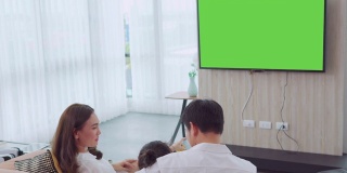 亚洲幸福家庭坐在家里的客厅看电视，一起度过宝贵的时间，频道到绿色屏幕显示器为您的信息