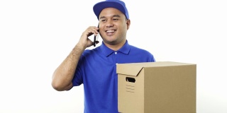微笑的年轻亚洲快递员身穿蓝色制服拿着包裹纸箱使用智能手机与客户在孤立的白色背景。4 k决议。