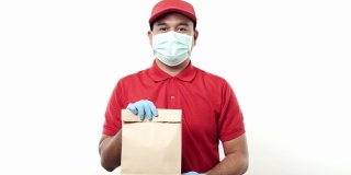 年轻的亚洲快递员穿着红色制服，戴着防护口罩和医用橡胶手套，在孤立的白色背景上给客户包裹纸袋。4 k决议。