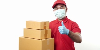 年轻的亚洲快递员身穿红色制服，戴着防护口罩和医用橡胶手套，拿着包裹纸板，竖起大拇指隔离。4 k决议。