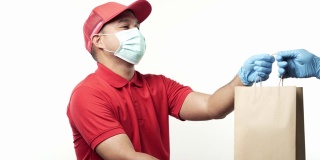 年轻的亚洲快递员穿着红色制服，戴着防护口罩和医用橡胶手套，在孤立的白色背景上给客户包裹纸袋。4 k决议。