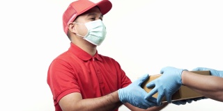 年轻的亚洲快递员身穿红色制服，戴着防护面具和医用橡胶手套，在孤立的白色背景下将包裹纸板交给客户。4 k决议。