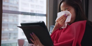 一个多病的年轻女子在用纸巾擤鼻子。坐在沙发上用笔记本电脑