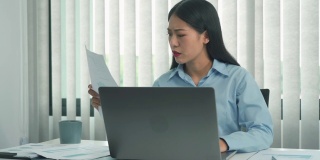 年轻的亚洲女性在办公室使用笔记本电脑时，对文件感到沮丧。