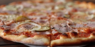 新鲜的圆形披萨，配上马苏里拉奶酪、意大利腊肠、黄瓜和洋葱，在棕色木桌上近距离从左到右旋转。披萨店的美味快餐背景