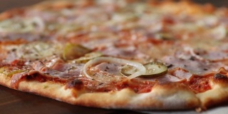 新鲜的圆披萨上有马苏里拉奶酪，意大利腊肠，黄瓜和洋葱，特写镜头从右到左在一个木制的棕色桌子上旋转。披萨店的美味快餐背景