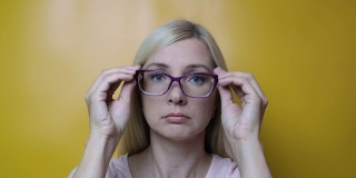 一个金发的中年女人戴上时尚的眼镜，对着黄色的背景微笑，近视，散光和眼科疾病的概念