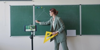 在线培训中，老师戴着眼镜，手里拿着尺子，在学校的黑板旁教数学课，用手机拍照
