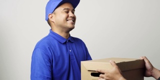 微笑的年轻亚洲快递员在蓝色制服给客户的包裹硬纸板和显示大拇指在孤立的白色背景。4 k决议。