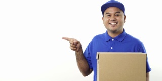 快乐年轻的亚洲快递员在蓝色制服拿着包裹硬纸板和指向空白的手指上的文字孤立的白色背景。4 k决议。