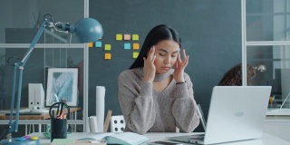 压力大的亚洲女性在办公室用笔记本电脑打字，患有偏头痛