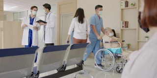 跟踪拍摄:两名医生在医院大堂或走廊与忙碌的护士、坐轮椅的老年患者、病人、带着医用口罩在接待处等候的亲属一起讨论、交谈。