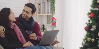 年轻的亚洲情侣情侣一起坐在沙发上。在以圣诞树为背景的房间里，用信用卡和笔记本电脑进行网上购物。4 k慢动作。