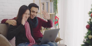 年轻的亚洲情侣情侣一起坐在沙发上。用笔记本电脑与家里装饰着圣诞树的房间里的家人和朋友进行视频通话。4 k慢动作。