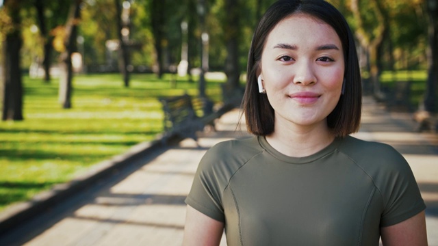 可爱的亚洲女人戴着无线耳机在城市公园对着摄像头微笑，跟踪拍摄