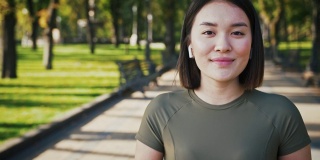 可爱的亚洲女人戴着无线耳机在城市公园对着摄像头微笑，跟踪拍摄