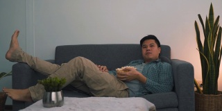 一名亚洲男子在晚上看电影时斜靠在沙发上吃宵夜。