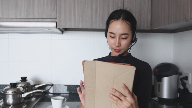 亚洲女性戴着耳机和使用数字平板电脑视频会议和会议，在家里的厨房喝咖啡